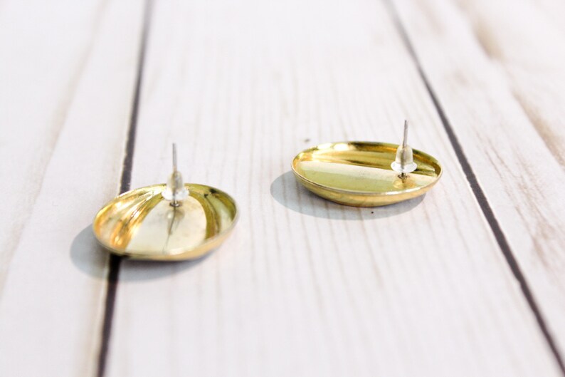 May include: Two gold-colored, oval-shaped stud earrings with silver posts. The earrings have a shiny, reflective surface. The earrings are displayed on a white wooden surface.