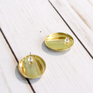 May include: Two gold-colored, round stud earrings with silver posts. The earrings have a shiny, metallic finish and are set against a light-colored, wooden surface. The earrings are simple and elegant.
