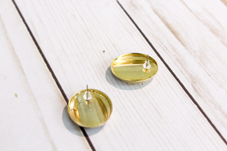 May include: Two gold-colored, round stud earrings with silver posts. The earrings have a shiny, metallic finish and are set against a light-colored, wooden surface. The earrings are simple and elegant.