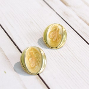 May include: A pair of vintage gold-tone clip-on earrings. Each earring has an oval shape with a cream-colored enamel center featuring a swirl design in shades of peach and gold. The earrings are set against a white wooden background.