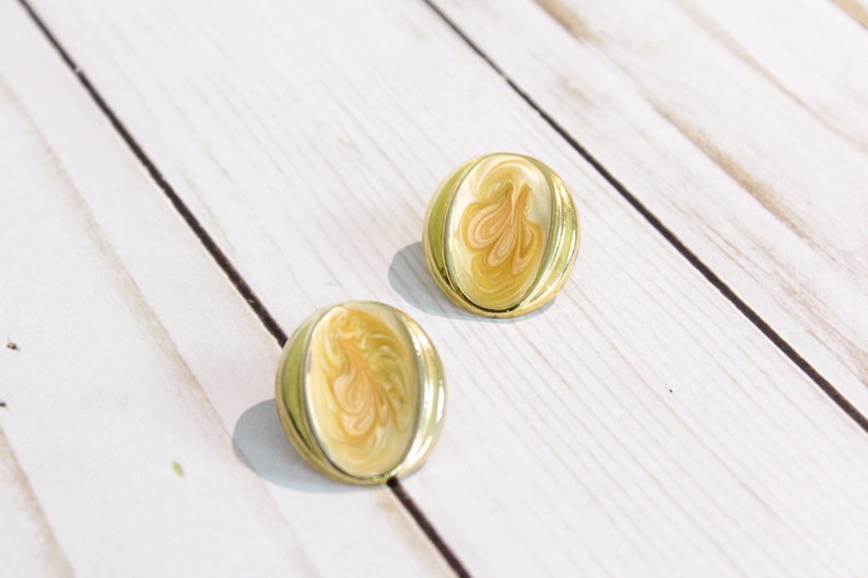 May include: A pair of vintage gold-tone clip-on earrings. Each earring has an oval shape with a cream-colored enamel center featuring a swirl design in shades of peach and gold. The earrings are set against a white wooden background.