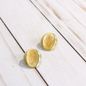 May include: A pair of gold-toned stud earrings. Each earring features a cream-colored, oval-shaped center with a swirling, abstract design in shades of peach and cream. The earrings are set against a white wooden background.