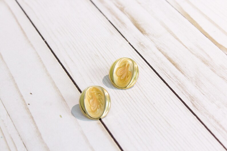 May include: A pair of gold-toned stud earrings. Each earring features a cream-colored, oval-shaped center with a swirling, abstract design in shades of peach and cream. The earrings are set against a white wooden background.