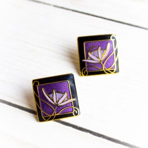 May include: A pair of square earrings with a floral design. Each earring features a purple flower on a black background, framed by gold-colored accents. The earrings are set against a white wooden surface.