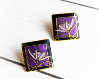 May include: A pair of square earrings with a floral design. Each earring features a purple flower on a black background, framed by gold-colored accents. The earrings are set against a white wooden surface.