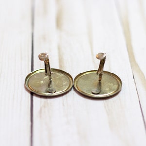 May include: A pair of gold-toned clip-on earrings with a round, flat design. The earrings have a simple, classic style.