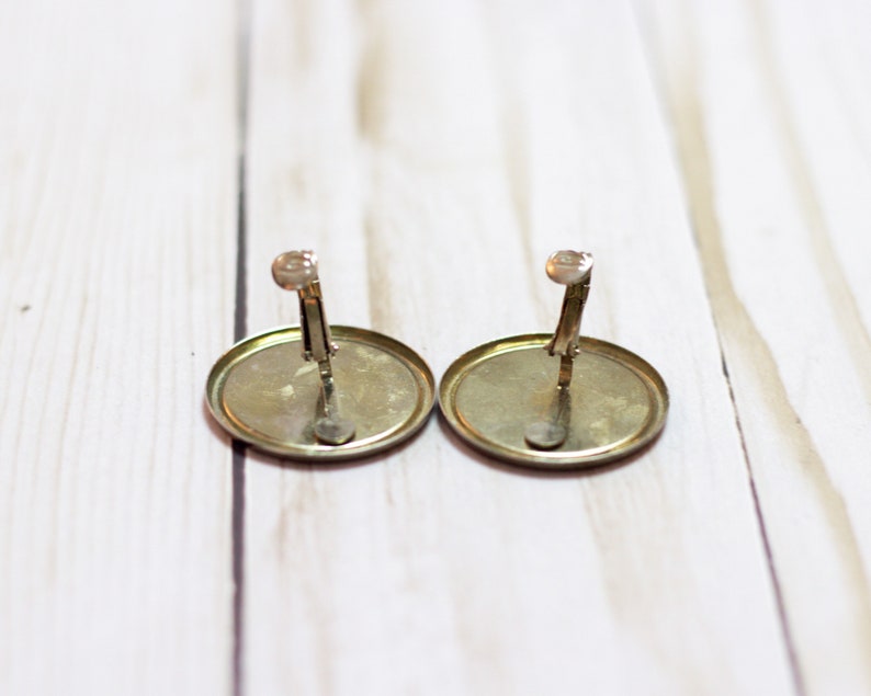 May include: A pair of gold-toned clip-on earrings with a round, flat design. The earrings have a simple, classic style.