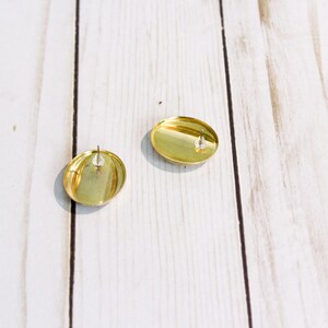 May include: Two gold-colored, oval-shaped stud earrings. The earrings have a shiny, metallic finish and a post backing. The earrings are displayed on a light-colored, wooden surface.