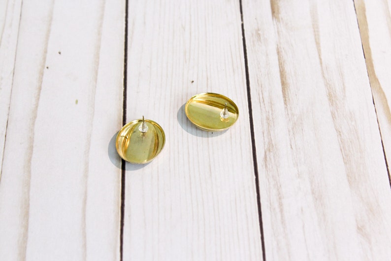 May include: Two gold-colored, oval-shaped stud earrings. The earrings have a shiny, metallic finish and a post backing. The earrings are displayed on a light-colored, wooden surface.