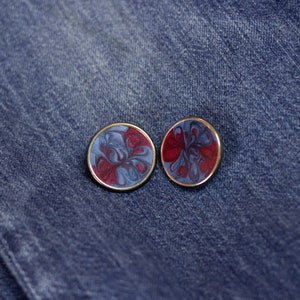 May include: Two round, gold-rimmed earrings with a swirling red and blue marbled design. The earrings are on a blue denim background.