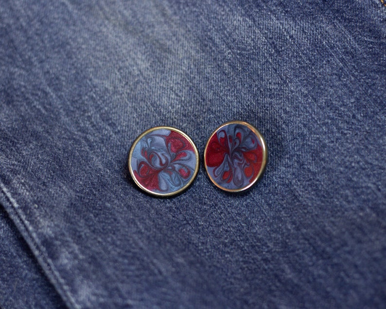 May include: Two round, gold-rimmed earrings with a swirling red and blue marbled design. The earrings are on a blue denim background.