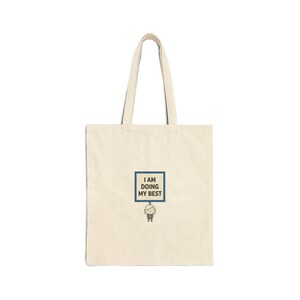 May include: Cream-colored tote bag with a rectangular graphic that reads "I AM DOING MY BEST" in a blue frame. A small cartoon figure is holding the sign. The bag has long handles.