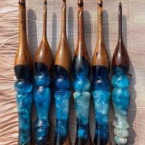 May include: Six handcrafted crochet hooks with wooden handles and blue resin bases. The wooden handles are a rich brown, and the resin sections feature swirling shades of blue and white. Each hook has a unique shape and design.