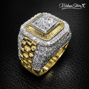 May include: A gold-toned ring with a square, diamond-studded center. The ring features multiple layers of diamonds and a textured band. The ring is set against a black background, and the brand name "Xurban StoreX" is visible in the top right corner.