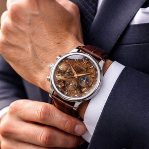 May include: A close-up of a Geneva watch with a brown leather band. The watch face is brown with silver accents and Roman numerals. The watch is on a wrist, and the person is wearing a dark suit.