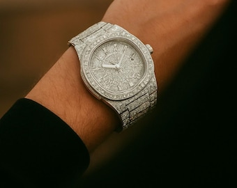 May include: A luxury wristwatch with a silver band and face, encrusted with numerous small, sparkling diamonds. The watch features a round face with Roman numeral hour markers and silver hands. The watch is on a person's wrist.