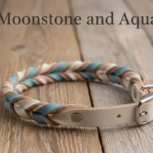 May include: A braided collar featuring alternating strands of light blue, cream, and tan leather. The collar includes a silver-toned metal buckle and a smooth leather strap section. The text Moonstone and Aqua is visible above the collar.