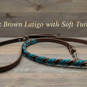Peut inclure: Une laisse pour chien en cuir latigo marron foncé avec une section tressée aux accents turquoise doux. La laisse comprend un mousqueton pivotant en métal et une poignée, avec le texte Dark Brown Latigo with Soft Turq visible au-dessus.