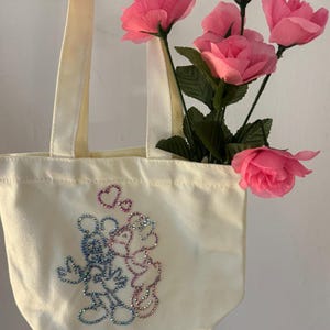 May include: A cream-colored tote bag with a rhinestone design of two cartoon mice embracing, with hearts above them. The bag has cream-colored handles and is filled with pink roses.