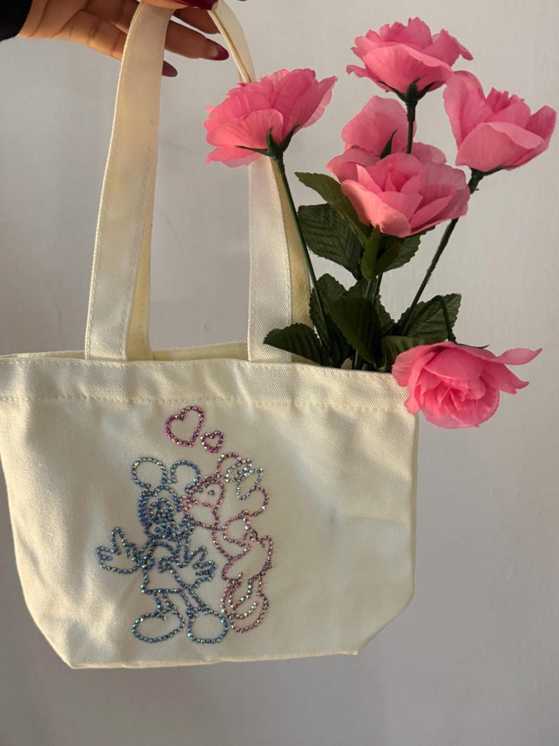 May include: A cream-colored tote bag with a rhinestone design of two cartoon mice embracing, with hearts above them. The bag has cream-colored handles and is filled with pink roses.