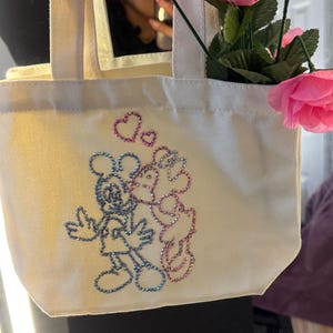 May include: Cream-colored tote bag featuring a glittery illustration of Mickey and Minnie Mouse kissing, with hearts above them. The bag is filled with pink roses. The bag has a rectangular shape with two handles.