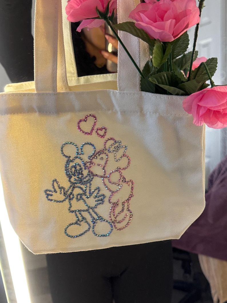 May include: Cream-colored tote bag featuring a glittery illustration of Mickey and Minnie Mouse kissing, with hearts above them. The bag is filled with pink roses. The bag has a rectangular shape with two handles.