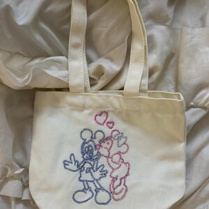 May include: Off-white canvas tote bag with two handles. The bag features an embroidered design of Mickey and Minnie Mouse kissing, with pink hearts above them. The characters are outlined in glittery blue and pink thread.
