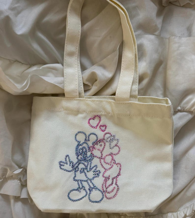 May include: Off-white canvas tote bag with two handles. The bag features an embroidered design of Mickey and Minnie Mouse kissing, with pink hearts above them. The characters are outlined in glittery blue and pink thread.