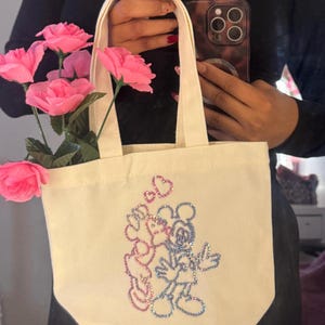 May include: Cream-colored tote bag featuring a sparkly Mickey and Minnie Mouse design. The bag has a rectangular shape with a flat bottom and two handles. Pink roses are placed inside the bag. The bag is held by a person.