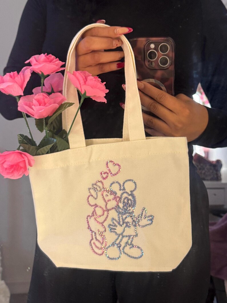 May include: Cream-colored tote bag featuring a sparkly Mickey and Minnie Mouse design. The bag has a rectangular shape with a flat bottom and two handles. Pink roses are placed inside the bag. The bag is held by a person.