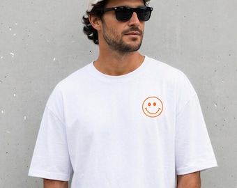 May include: White t-shirt with a small orange smiley face graphic. The shirt is a relaxed fit and is being worn with a baseball cap and sunglasses. The background is a plain gray wall.
