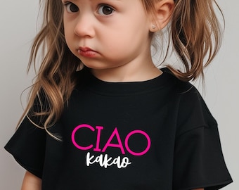 May include: A black t-shirt with the words "CIAO Kakao" in pink and white lettering. The t-shirt is worn by a young child with brown hair styled in pigtails. The child has a slightly sad expression.