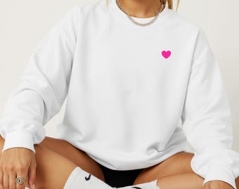 May include: White sweatshirt with a small, bright pink heart embroidered on the left chest. The long-sleeved top has a crew neck and a relaxed fit. The model is wearing white socks and sneakers.