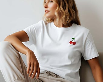 May include: White t-shirt with a small embroidered cherry design on the chest. The shirt is paired with beige sweatpants and white sneakers. The person is sitting on a gray couch.