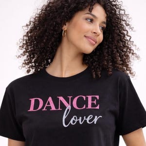 Könnte beinhalten: Schwarzes Crop-Top mit dem Text "DANCE lover" in rosa Glitzer und weißer Schrift. Das kurzärmelige Shirt ist ein lässiger Stil.
