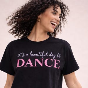 Könnte beinhalten: Schwarzes, kurzes T-Shirt mit dem Text "it's a beautiful day to DANCE" in Weiß und Pink. Das Shirt hat einen Rundhalsausschnitt und kurze Ärmel. Das Model lächelt und trägt goldene Creolen.