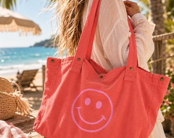 May include: A coral-colored canvas tote bag featuring a large, light pink smiley face graphic on the front. The bag has two wide shoulder straps attached with metal rivets and a snap button closure at the top opening for securing items inside.