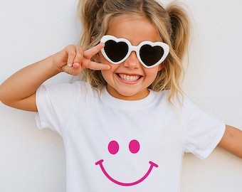 May include: A young person wearing white heart-shaped sunglasses and a white t-shirt with a pink smiley face graphic. The person is smiling and making a peace sign with their fingers.