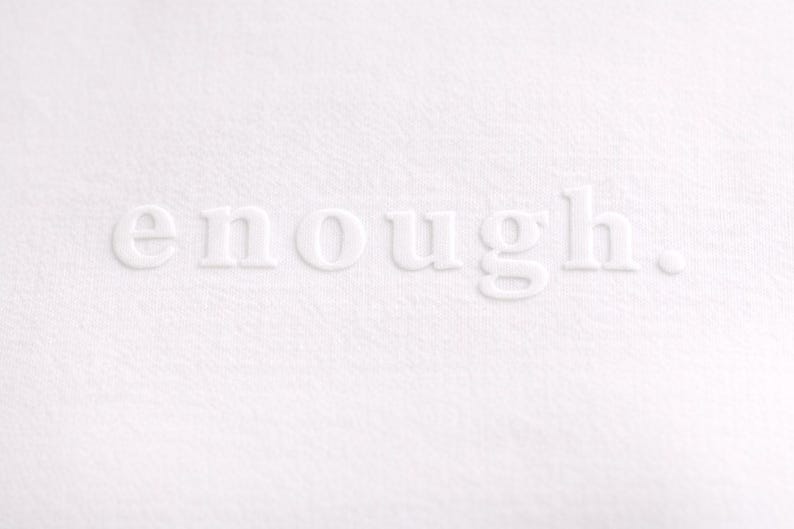 May include: A close-up of a white textured surface with the word "enough" embossed in white. The word is centered and the period is visible. The background is a solid white color.
