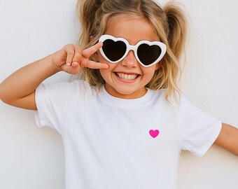 May include: A young person wearing white heart-shaped sunglasses and a white t-shirt with a small pink heart. The person is smiling and making a peace sign with their fingers. The background is a plain white wall.