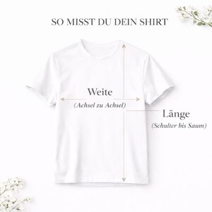 May include: A white t-shirt with text and arrows indicating how to measure the shirt. The text reads "SO MISST DU DEIN SHIRT", "Weite (Achsel zu Achsel)", and "L&auml;nge (Schulter bis Saum)". The shirt is displayed on a light background with floral accents.