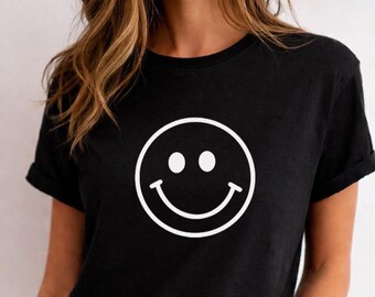 May include: Black t-shirt featuring a large white smiley face graphic. The design includes two white dots for eyes and a curved line for the mouth. The short-sleeved shirt has rolled-up sleeves.
