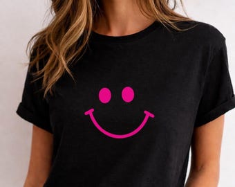 May include: Black t-shirt featuring a large, bright pink smiley face graphic. The design includes two pink circles for eyes and a curved pink line for the mouth. The shirt has short sleeves and a crew neck.