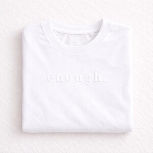 May include: A folded white t-shirt with the word "enough" subtly embossed across the front. The shirt has a crew neck and is made of a soft, likely cotton, material. The minimalist design and neutral color make it versatile.