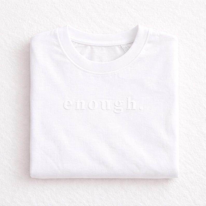 May include: A folded white t-shirt with the word "enough" subtly embossed across the front. The shirt has a crew neck and is made of a soft, likely cotton, material. The minimalist design and neutral color make it versatile.