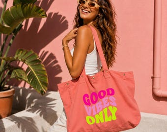 May include: A coral-colored canvas tote bag featuring the text "GOOD VIBES ONLY" printed in wavy, stacked lines of bright pink, light pink, and lime green. The bag has two shoulder straps, metal rivets at the base of the straps, and a wide opening.