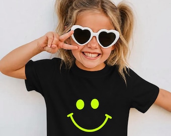 May include: A child wearing a black t-shirt with a neon yellow smiley face graphic. The child is also wearing white heart-shaped sunglasses and making a peace sign with their fingers.