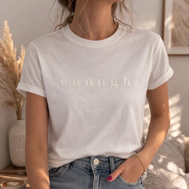 May include: White t-shirt with rolled sleeves and the word "enough" embroidered in a matching white thread. The shirt is paired with blue jeans. A gold bracelet is visible on the wrist.