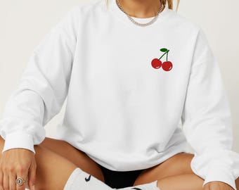 May include: White sweatshirt with a crew neck and long sleeves. The sweatshirt features a small embroidered design of two red cherries with a green stem on the upper left chest. The model is wearing black shorts and white sneakers.