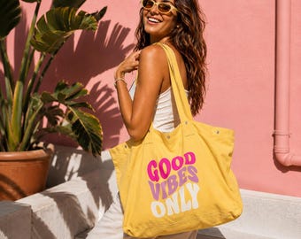 May include: A yellow canvas tote bag featuring the text "GOOD VIBES ONLY" printed in stacked, wavy lettering. The word "GOOD" is in bright pink, "VIBES" is in light purple, and "ONLY" is in white. The bag has two shoulder straps and metal rivets.